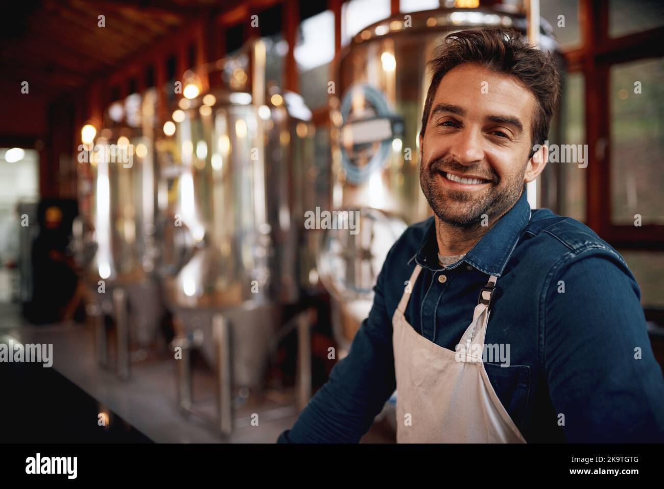 Doing what he does best. Portrait of a cheerful young barman leaning on the bar counter waiting for customers to serve inside of a beer brewery. Stock Photo