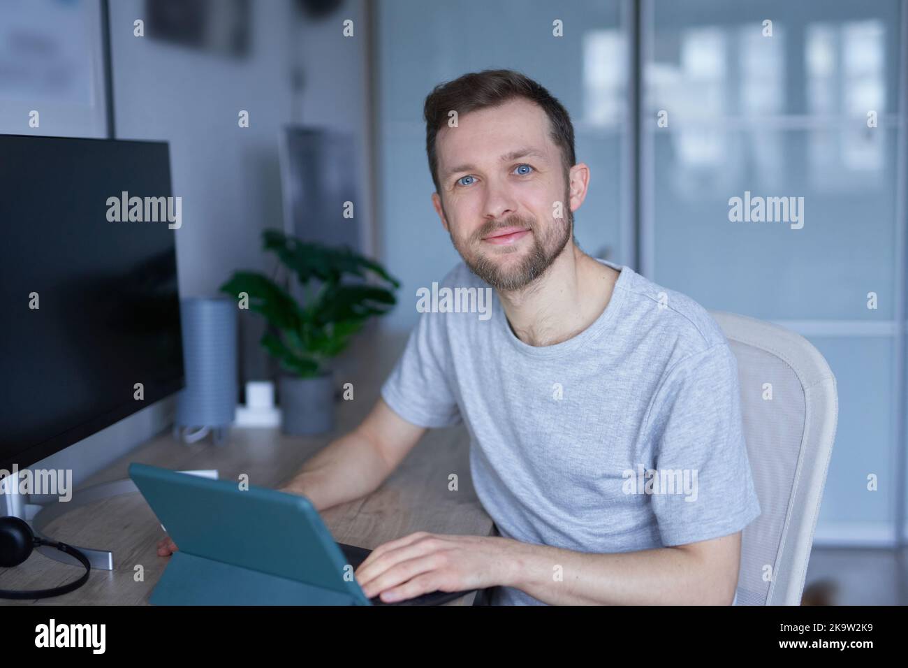 Smart tech concept. Attractive bearded male sitting in home office using tablet. Close-up of portrait of young handsome man in grey shirt and looking in camera, positive emotion. High quality photo Stock Photo