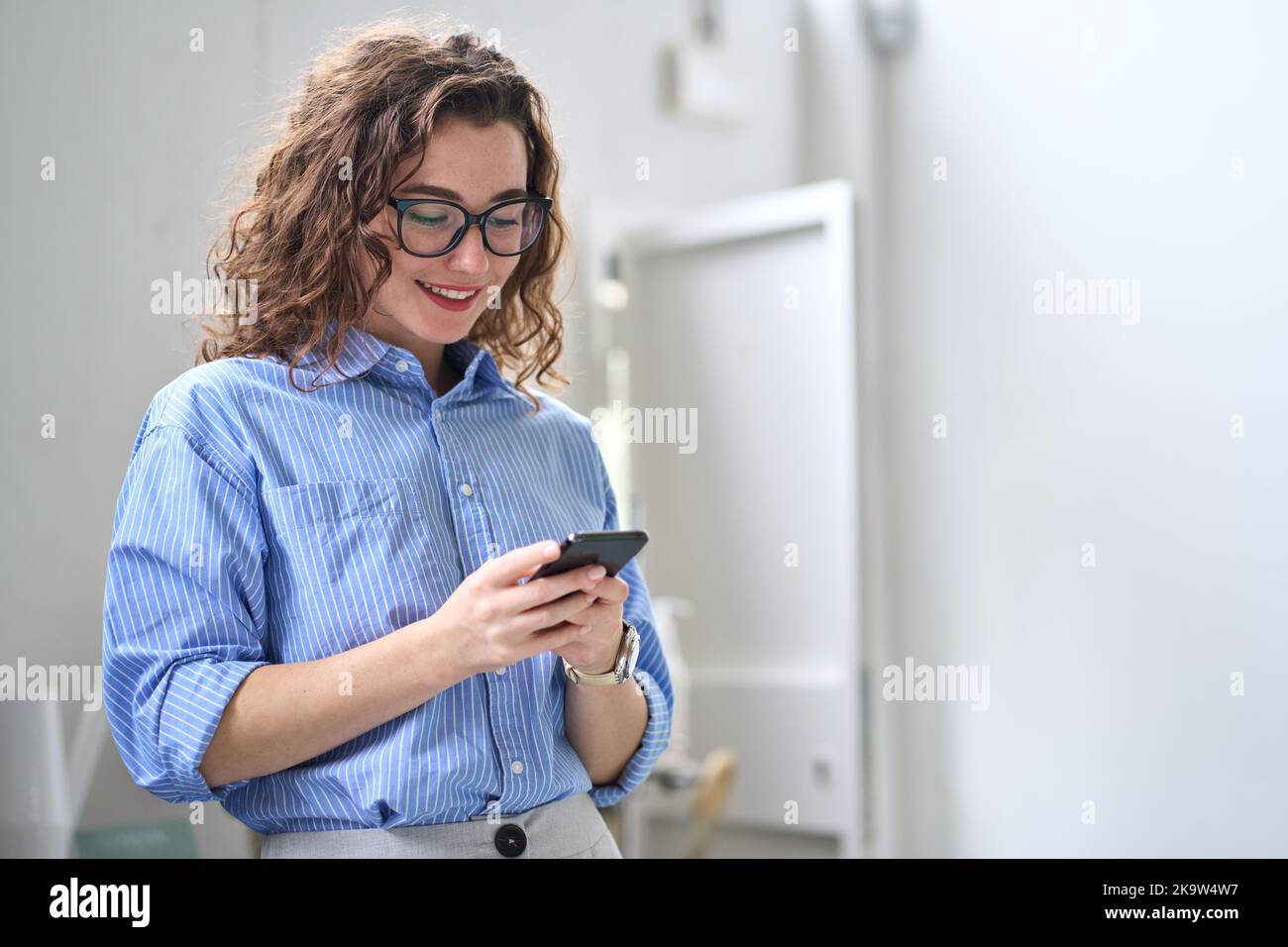 Smiling young business woman using cellphone standing in office using mobile. Stock Photo