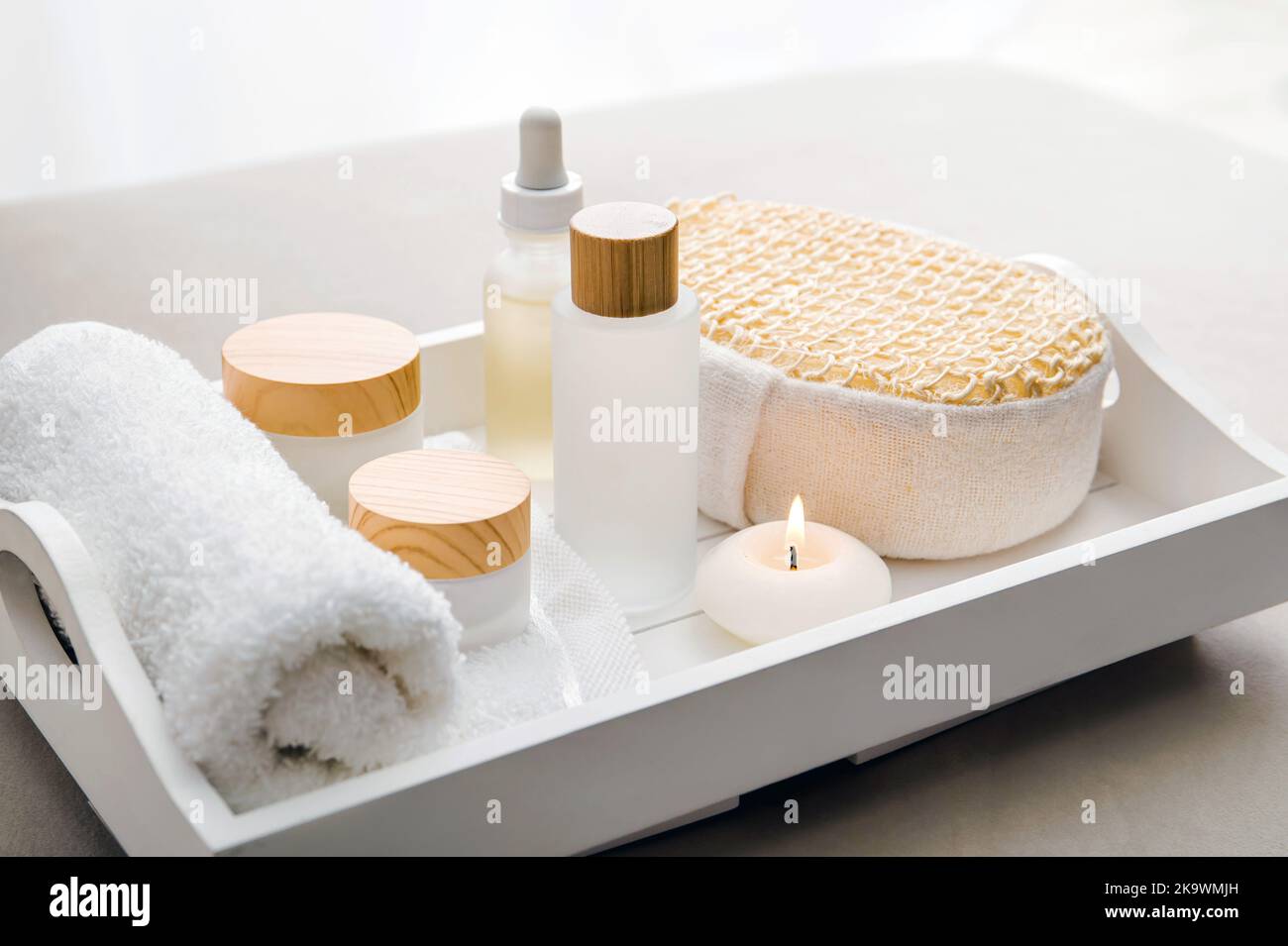 Various spa products on white tray, luxury spa relaxation concept. Massage oil, moisturizing creams, day cream, bath sponge, round candle burning. Stock Photo