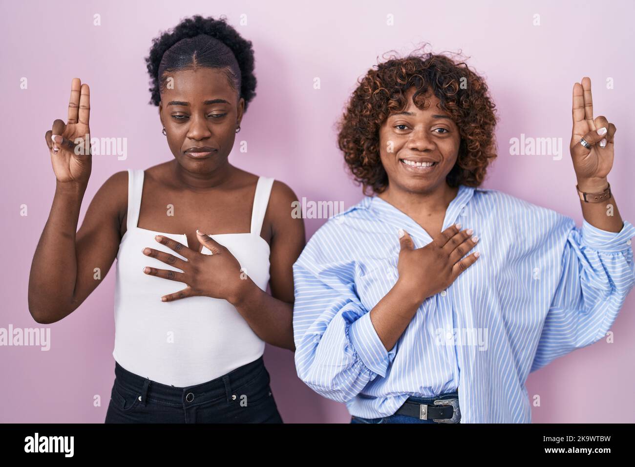 Two african women standing over pink background smiling swearing with ...