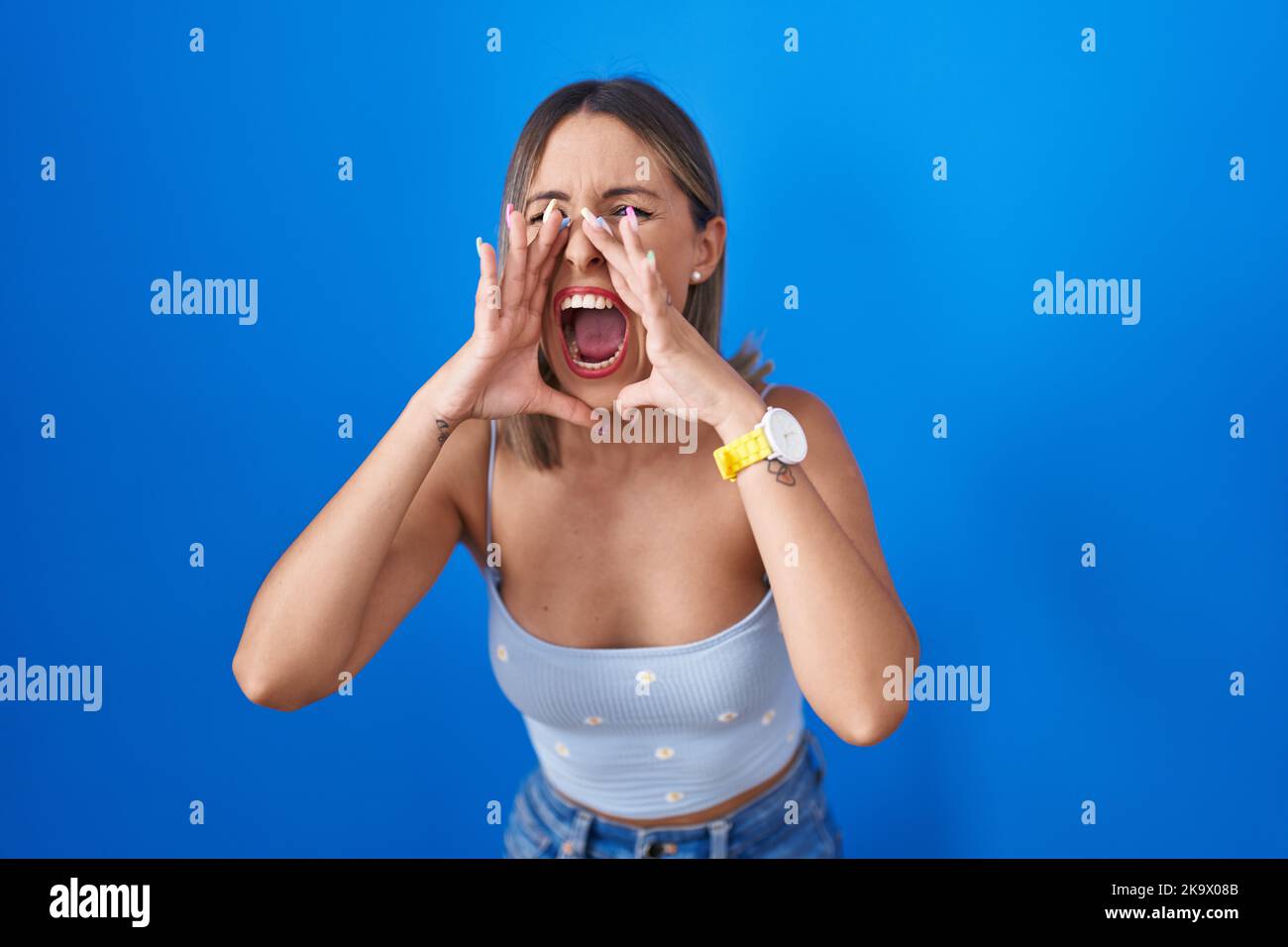 Young woman standing over blue background shouting angry out loud with ...