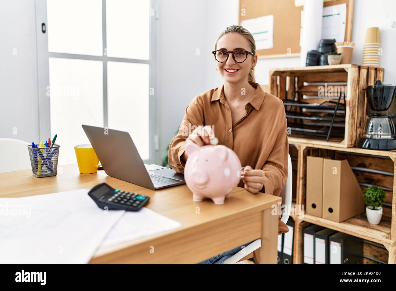 Young blonde woman at the office putting coin into piggy bank looking ...