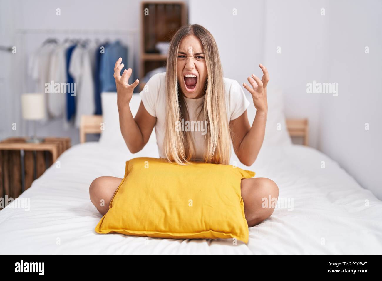 Young blonde woman sitting on the bed with pillow at home crazy and mad ...