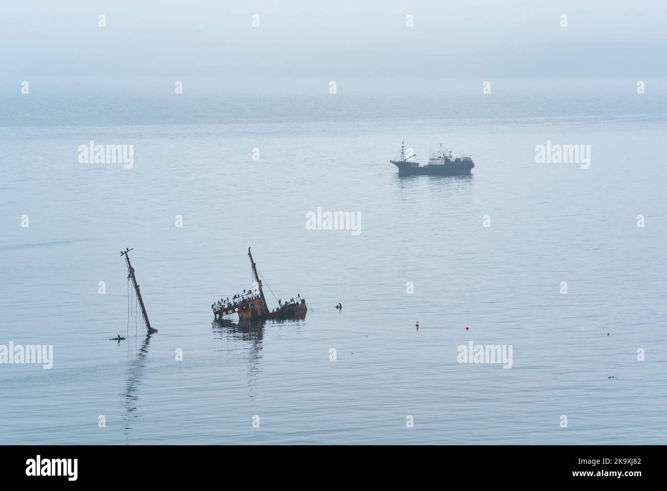fishing ship and sunken shipwreck at sea against the background of a distant foggy coast Stock Photo