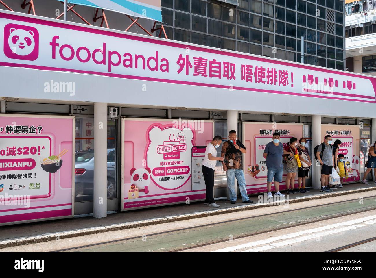 Commuters wait at a tram stop station displaying a commercial advertisement of the delivery take