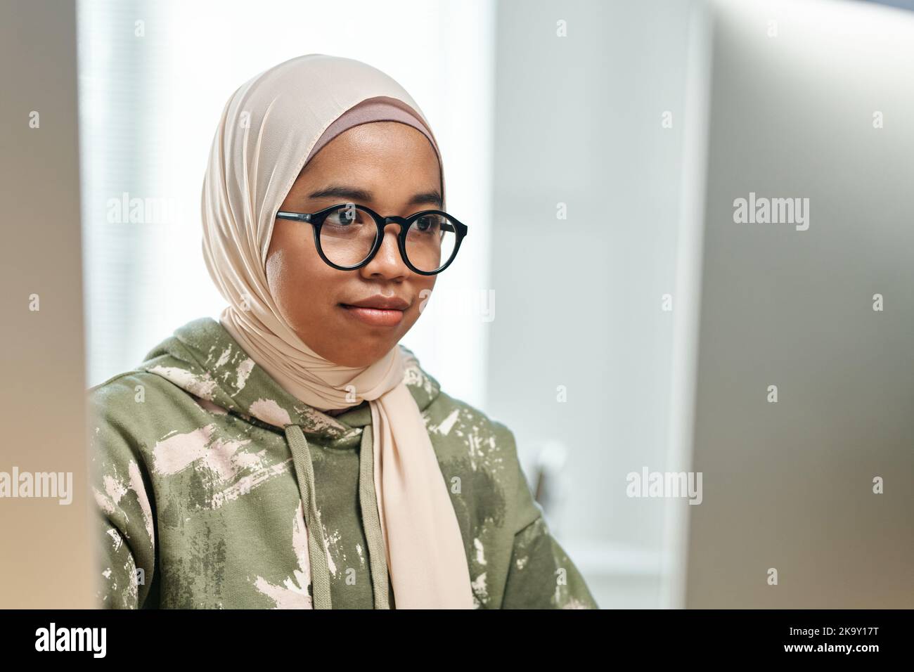 Young Muslim black woman in hijab and eyeglasses sitting in front of computer screen in office while developing new software Stock Photo