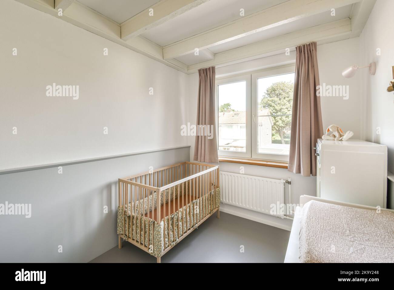 Interior of white room with baby crib near window with chest of drawers