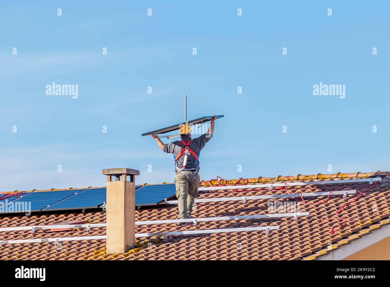 A technician lifts a solar panel over his head as he prepares to mount it on the roof of a red-tiled house Stock Photo