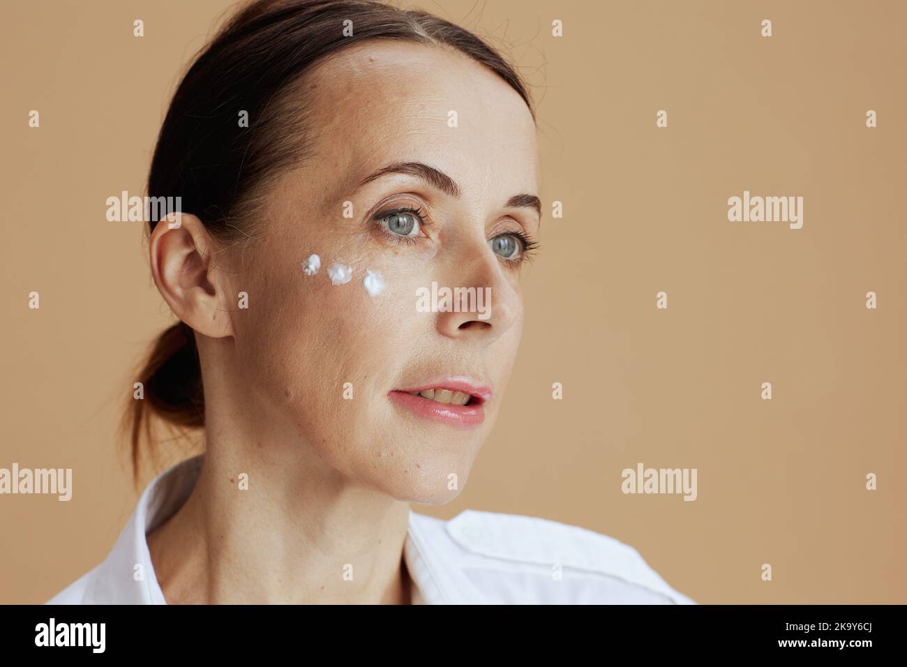 modern 40 years old woman with eye cream on face isolated on beige