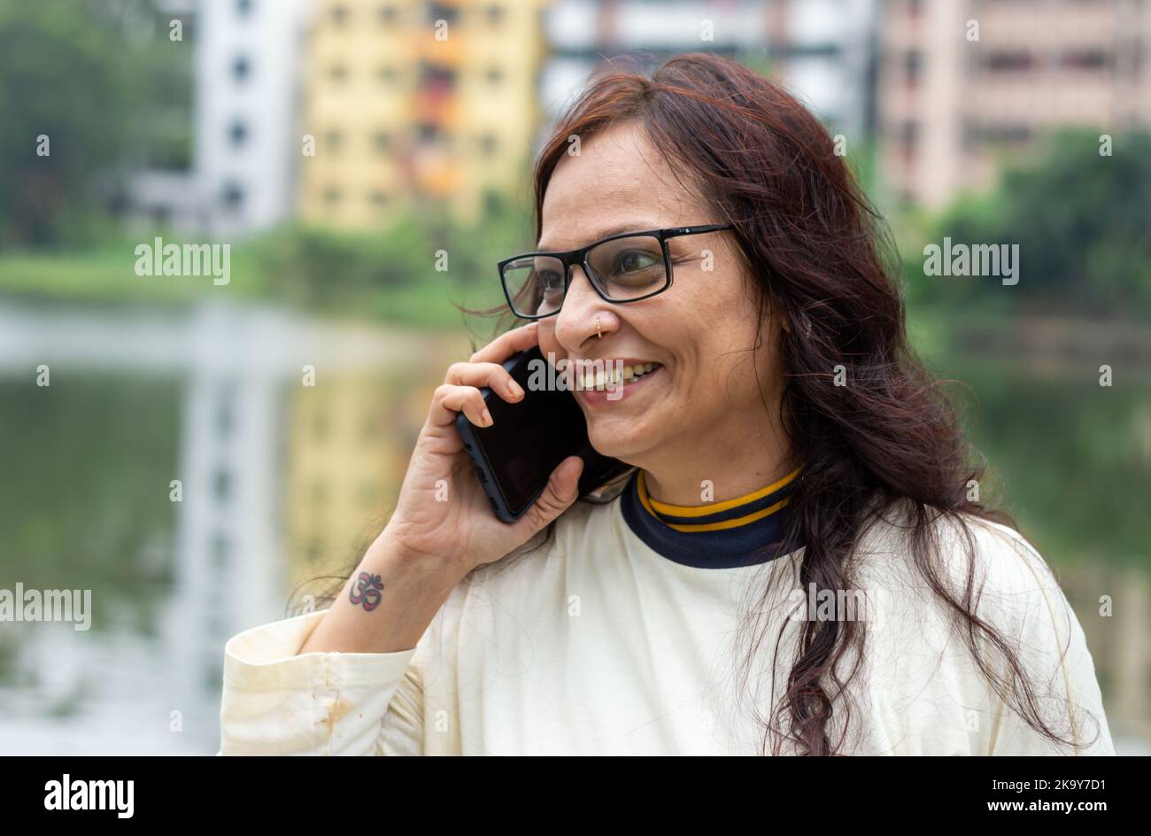 Mid Adult Smiling Woman with long brown hair Speaking on mobile phone outdoors. Closeup. She is wearing White T-shirt and specs. Front view. Head and Stock Photo