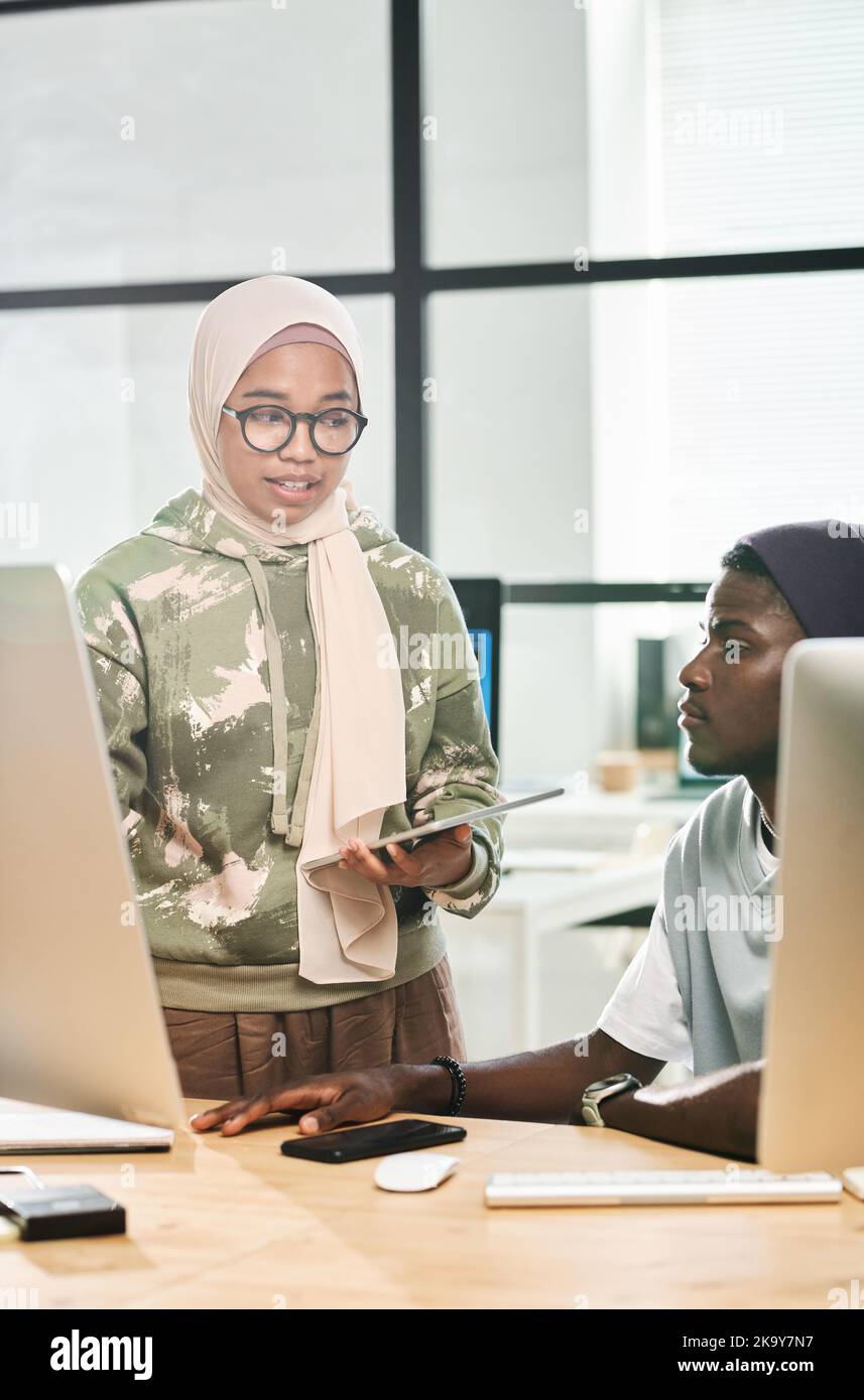 Young Muslim businesswoman in hijab explaining somthing to African American male colleague sitting between two monitors Stock Photo