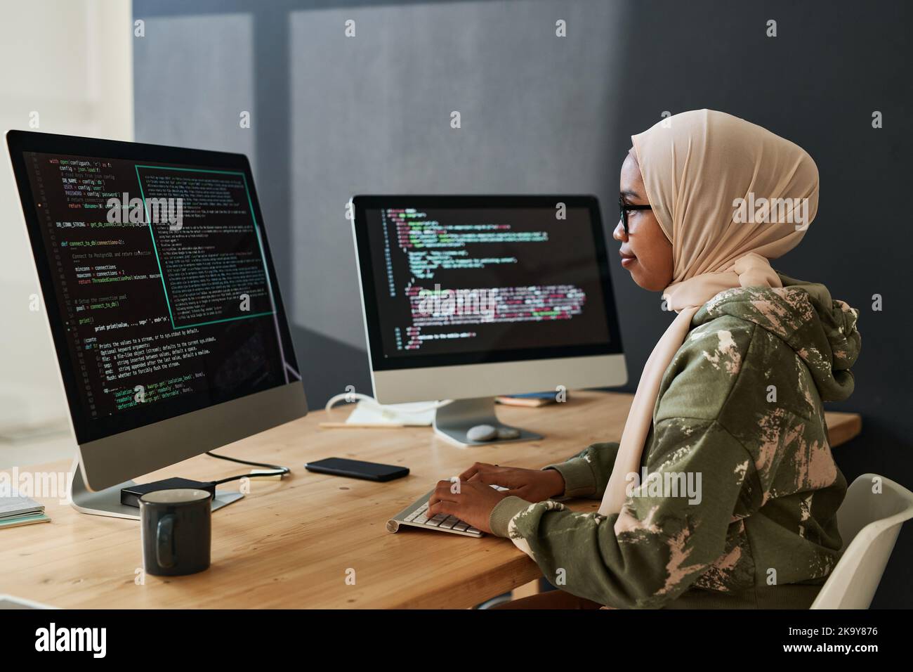 Side view of young Muslim female programmer looking through coded information on computer screen while sitting by workplace Stock Photo