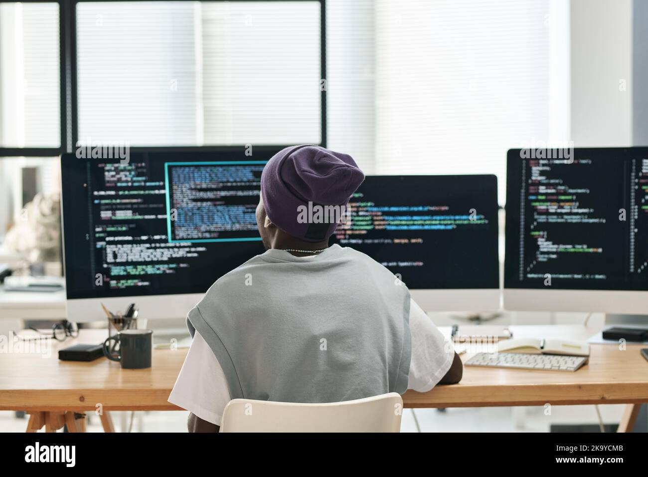 Back view of young software developer sitting by workplace in front of three computer monitors with coded data on screens Stock Photo