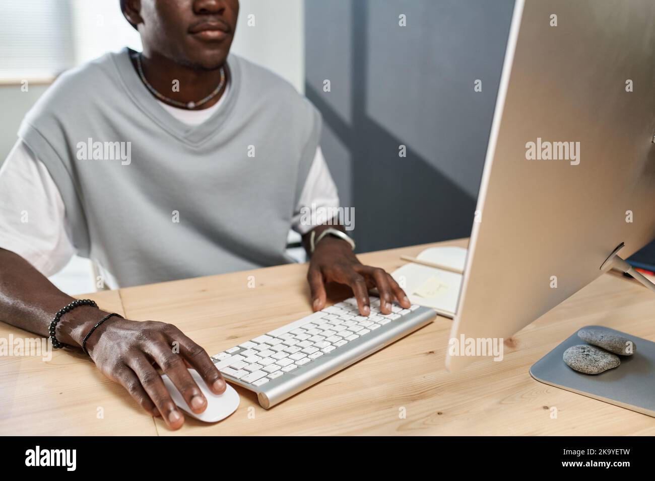 Close-up of young black man in t-shirt clicking on mouse and pressing button of computer keyboard while working in front of monitor Stock Photo
