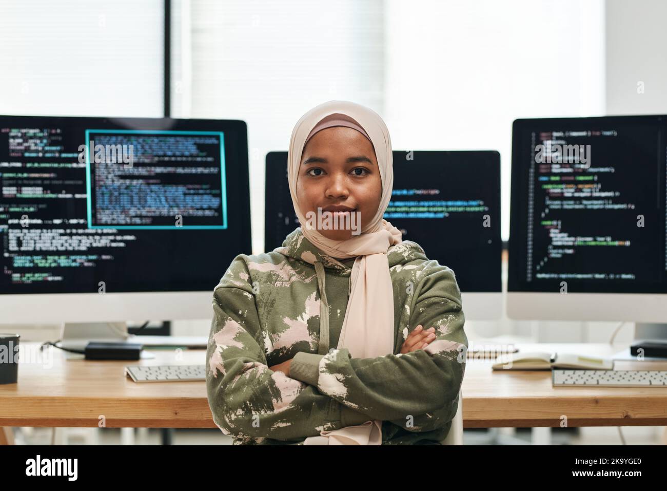 Young successful IT engineer in hijab looking at camera against workplace with computer monitors and coded data on their screens Stock Photo