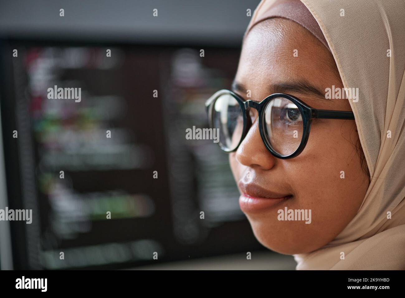 Close-up of face of young confident Muslim intern in hijab and eyeglasses standing in front of camera against computer screen with data Stock Photo