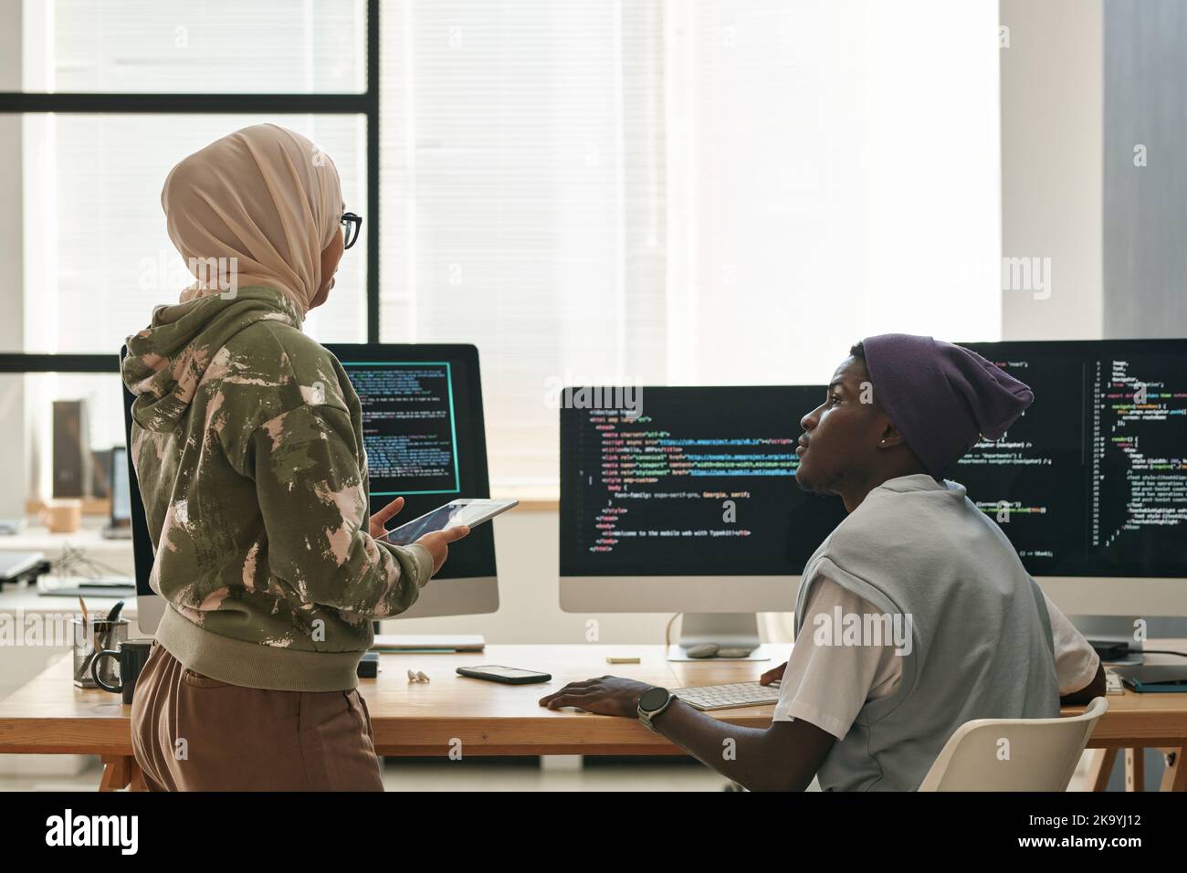 Two young intercultural IT engineers discussing ways of decoding data while Muslim woman in hijab standing in front of computer screens Stock Photo