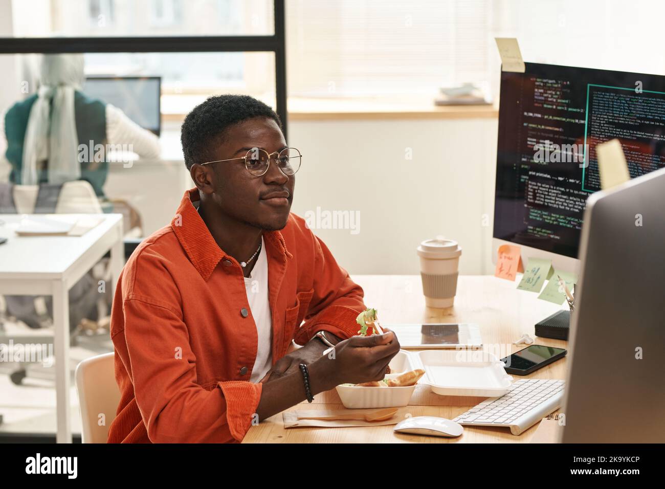 Young African American IT engineer looking at computer screen while having fresh vegetable salad from eco package for lunch Stock Photo