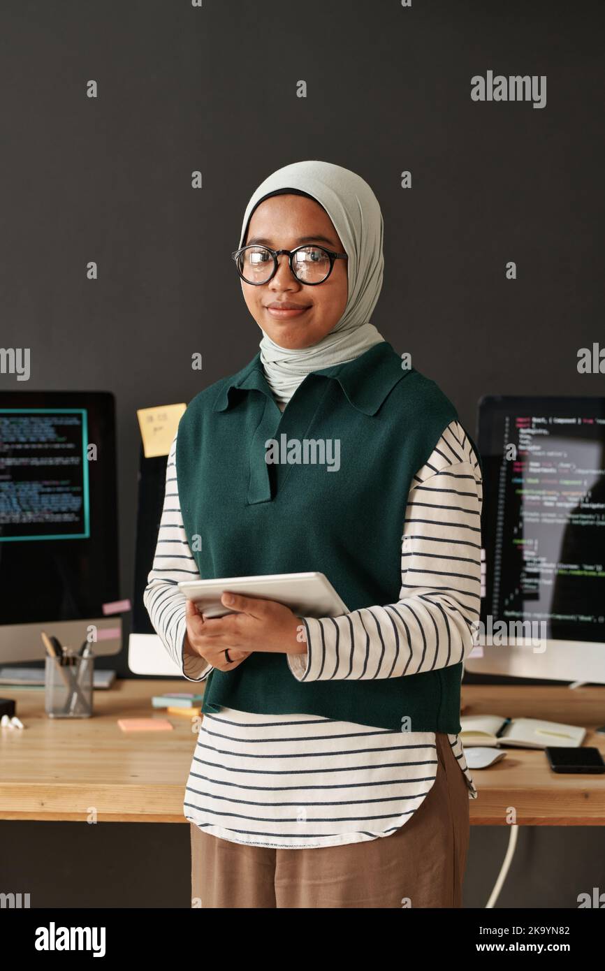 Young successful Muslim businesswoman or intern with tablet in hands looking at camera while standing by workplace with computers Stock Photo