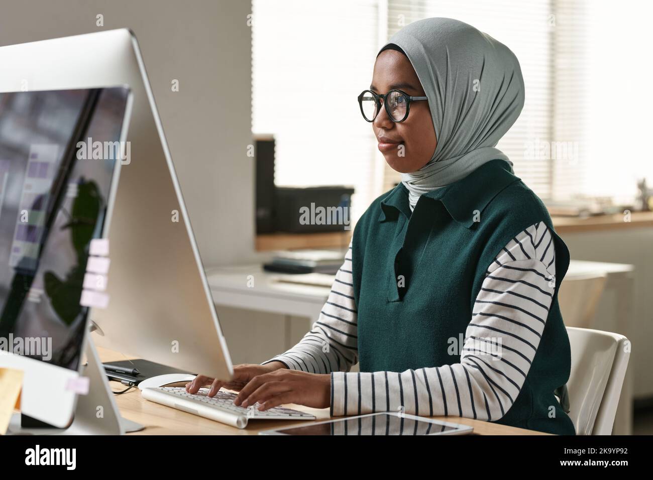 Young confident Muslim female intern or designer in eyeglasses, hijab and casualwear networking in front of computer monitor Stock Photo