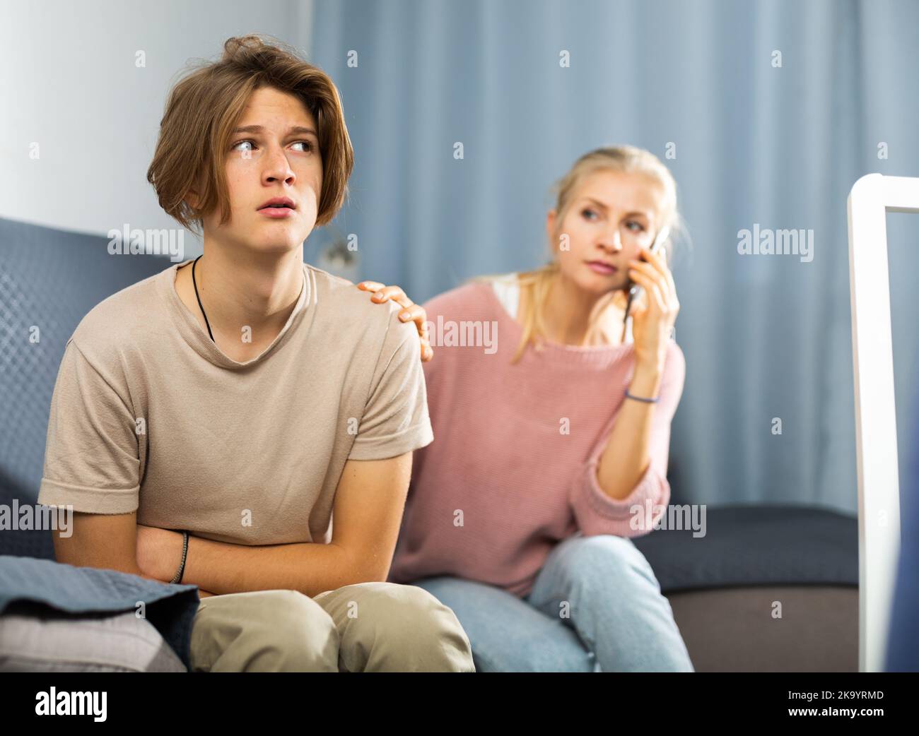 Teenager with stomach ache sitting at home while worried mom calling doctor Stock Photo