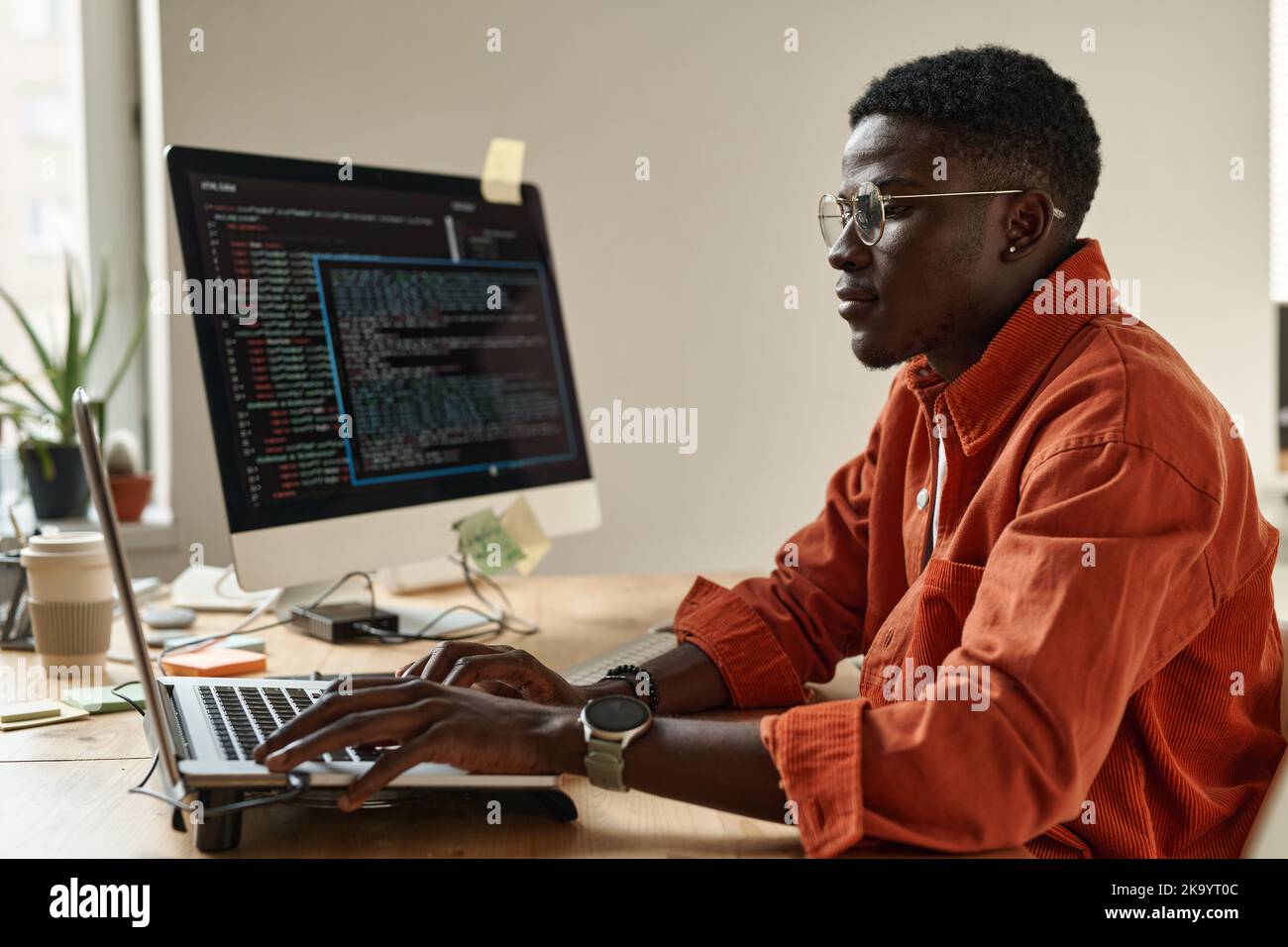 Side view of young black man in smart casualwear and eyeglasses decoding data while sitting by workplace in front of laptop Stock Photo