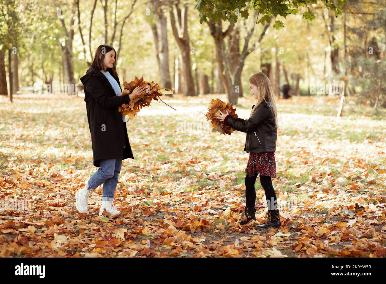 Cheerful young woman and little girl throw up bunch of dry yellow leaves. Happy mother and daughter in casual black outfits have fun and play in Stock Photo