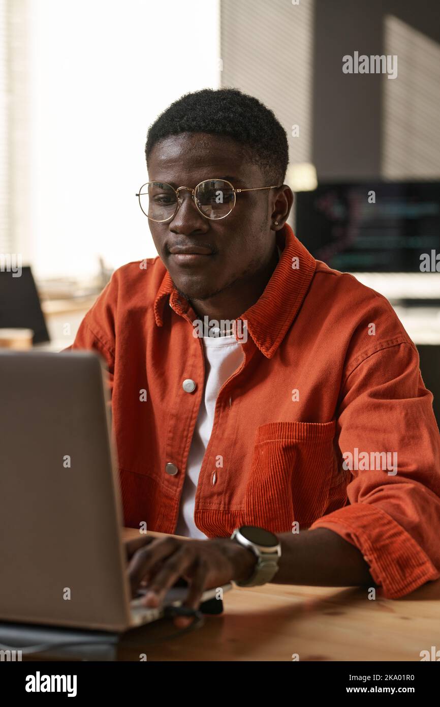 Serious young IT support engineer in casualwear and eyeglasses sitting by workplace in front of laptop and networking or decoding data Stock Photo
