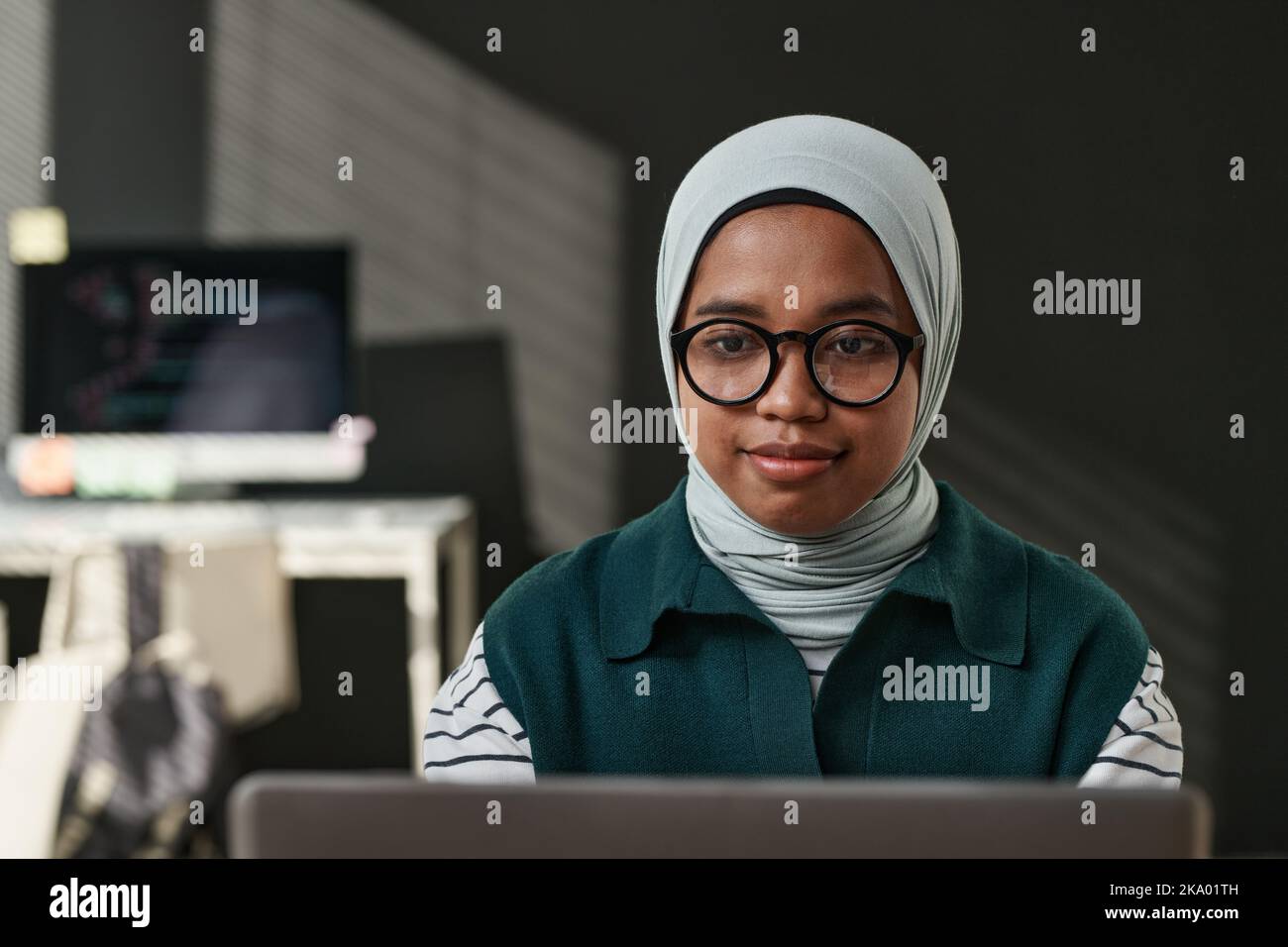 Young IT support engineer in casualwear, eyeglasses and hijab sitting in front of computer monitor and networking in office Stock Photo