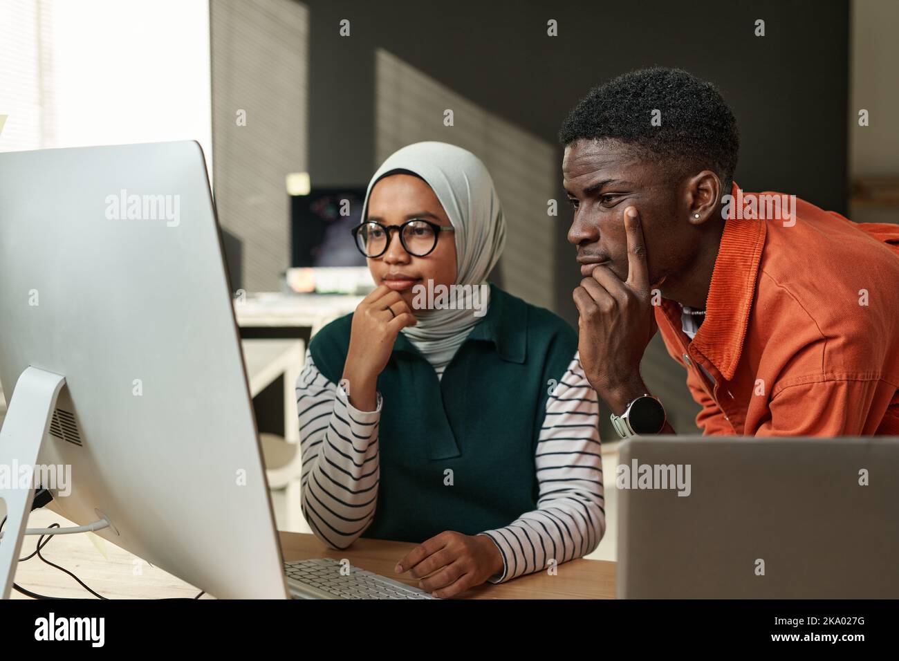 Two young serious intercultural IT engineers looking at data on computer screen or waiting for web page uploading at meeting in office Stock Photo