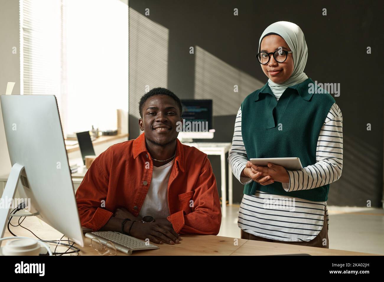 Two young software developers looking at camera while black man sitting by desk and Muslim woman with tablet standing next to him Stock Photo