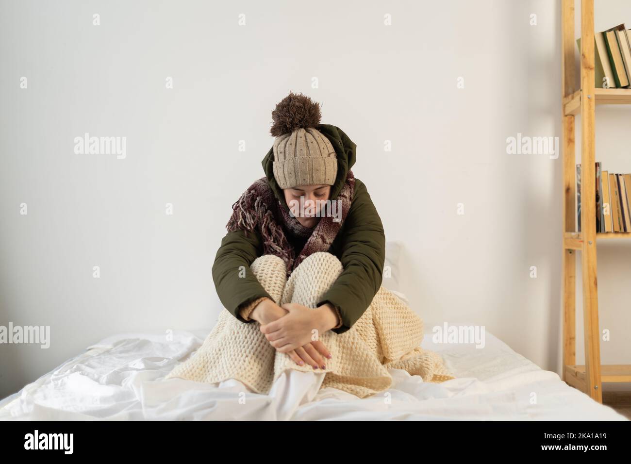 Angry young woman warmly dressed in a cold house sits on the bed feeling cold inside the house Stock Photo