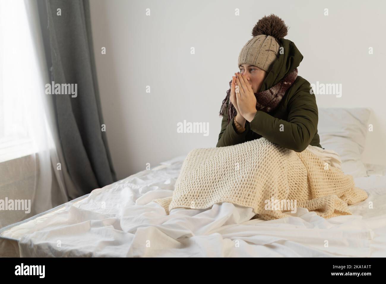 Sad female sit on bed at freezing cooled flat in warm cap and blanket shiver tremble with cold. Unhappy young woman at home feel bad suffer of heating Stock Photo