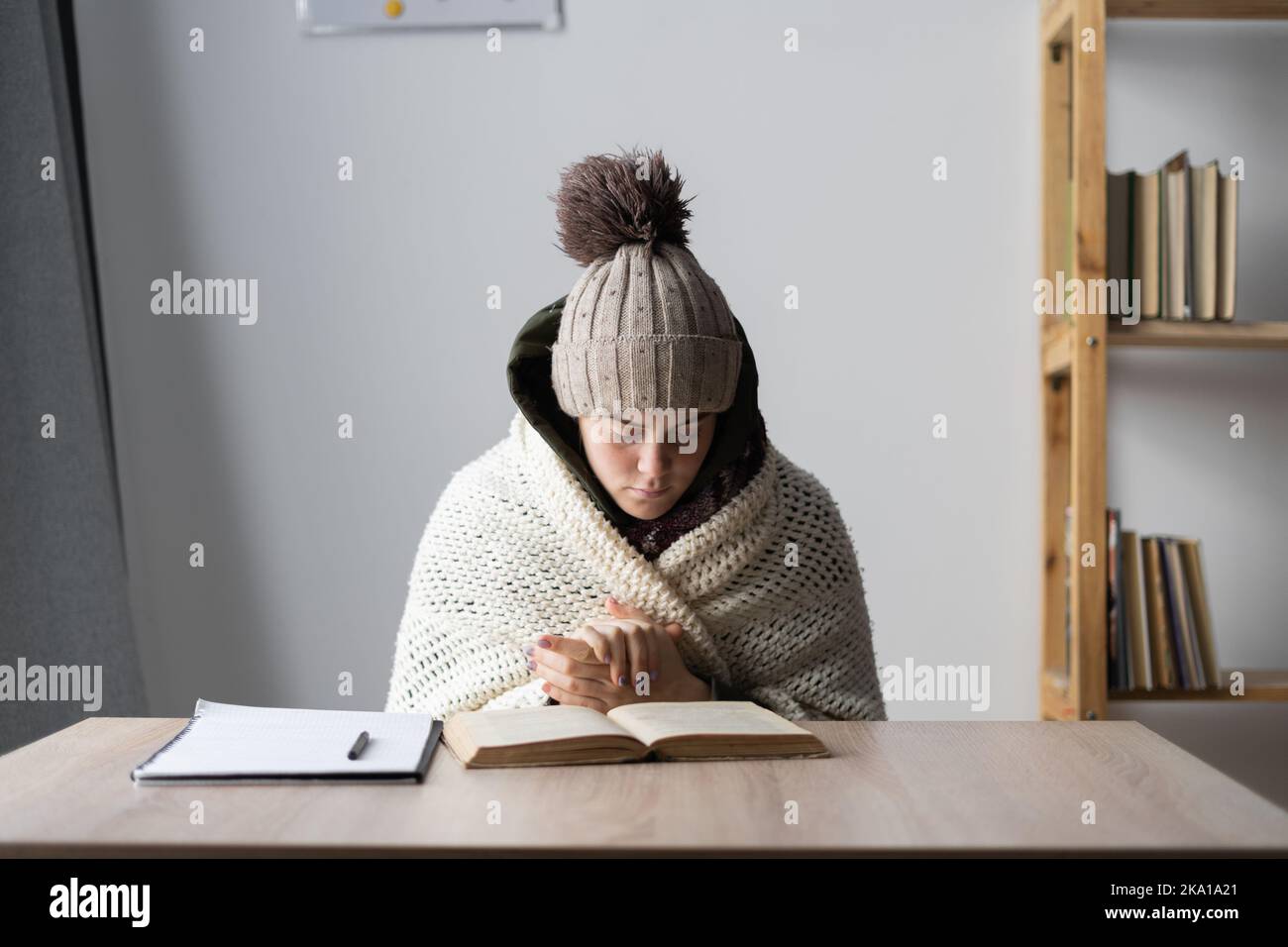 Cold freezing angry student sitting at the table at home. A young girl is studying suffering from cold without heating at home. No heating and energy Stock Photo