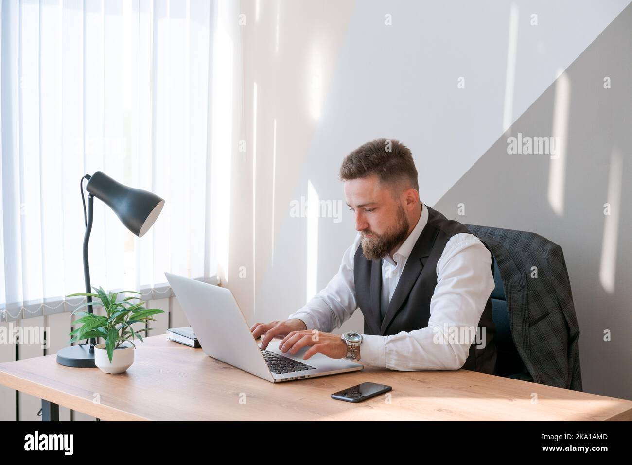 Caucasian man sits at table near laptop in office and looks at screen. Serious and thoughtful bearded boss or employee working at his workplace and feeling interested or analyzing the results. Stock Photo