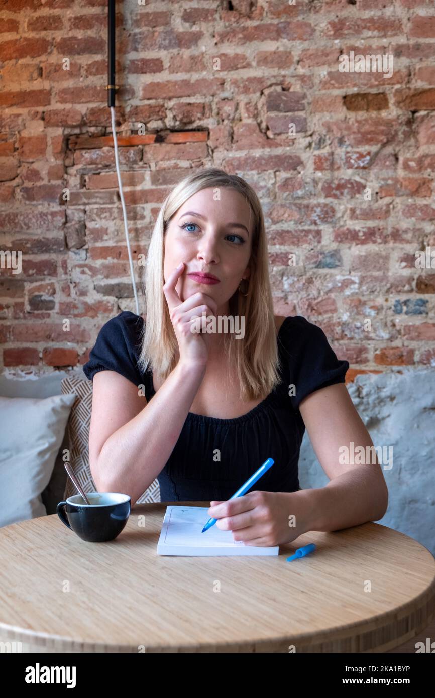 Young woman pondering and thinking about what to write down. Writer's block, future plans and goals in own diary resting in cozy cafeteria with cup of tasty cappuccino.Smiling hipster girl making getting good idea while writing in notepad. High quality photo Stock Photo