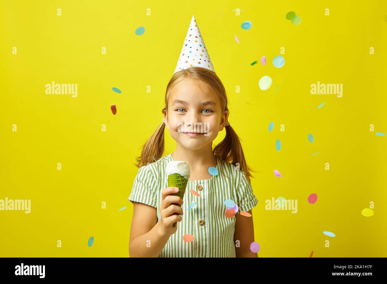 Happy little girl in a birthday hat, eating ice cream, it pours confetti, portrait on yellow isolated. Stock Photo