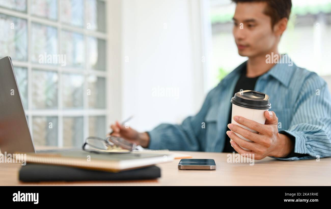 selective focus hand image, Handsome millennial Asian businessman in casual clothes holding a coffee cup while working on laptop. Stock Photo