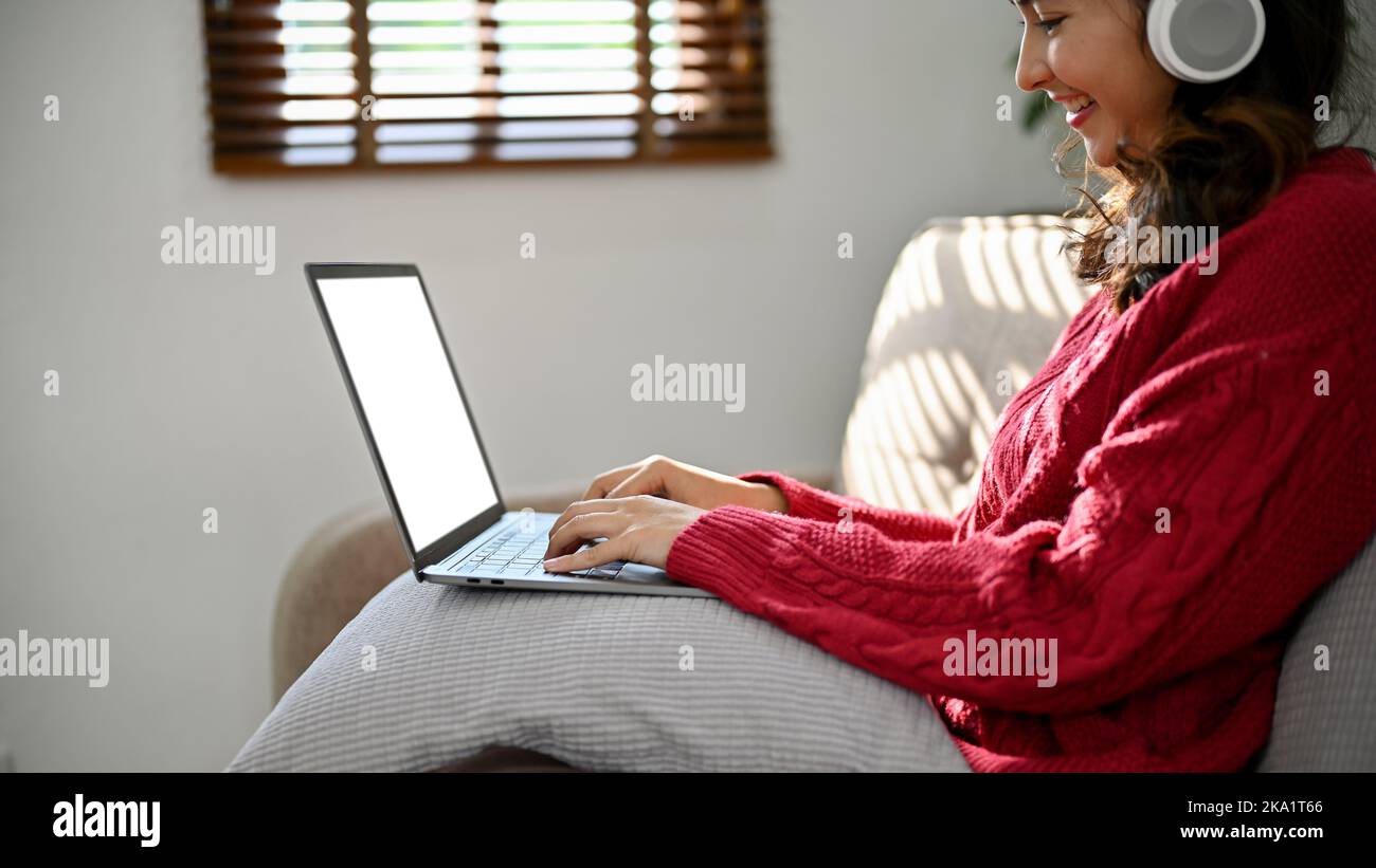 Beautiful young Asian female in red sweater wearing headphones, using laptop to manage her tasks or research online information while sitting on a cou Stock Photo