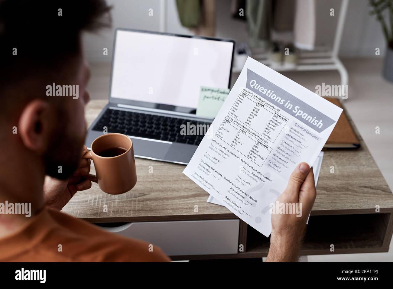 Close-up of young man with cup of tea and paper with questions in Spanish sitting by workplace and looking through assignment points Stock Photo