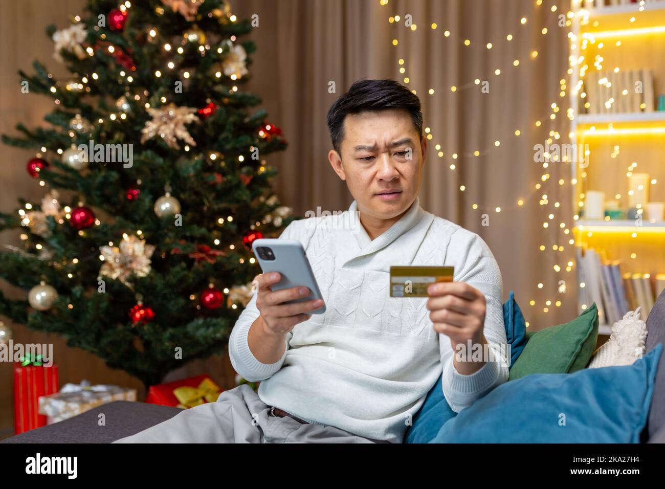 Worried and serious young Asian man sitting on sofa near Christmas tree at home. Holds phone and credit card in hands, can't pay for holiday online purchases, can't place an order. Stock Photo