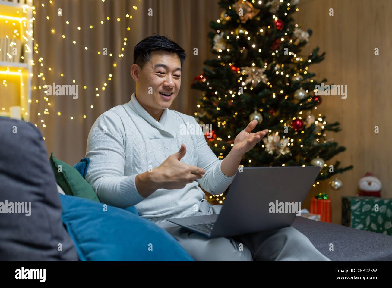 Quarantine for holidays. Young handsome Asian man sitting at home on sofa alone on New Year near Christmas tree. Holds a laptop, talks to family, friends via online video call. Stock Photo