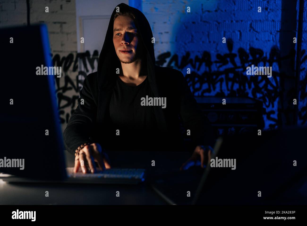 Portrait of hidden hacker man wearing sweatshirt with hood engaged in hacking into security systems, sitting in dark basement room with blue neon Stock Photo
