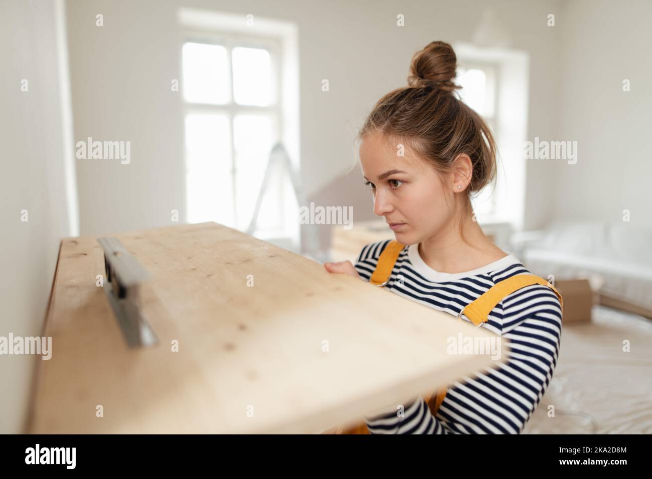 Young woman remaking her apartment, measuring wall with spirit level and hanging shelf. Stock Photo