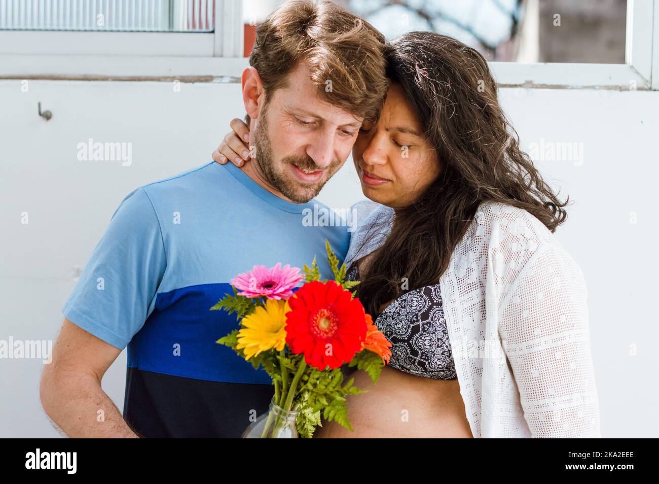 pregnant young couple argentinian man and brazilian woman are standing at home embracing each other holding flowers and looking at the flowers, they a Stock Photo