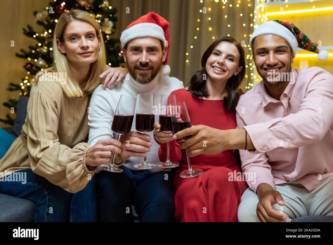 Portrait of a group of diverse guests at home for the new year, friends celebrating Christmas looking at the camera and smiling holding glasses of wine, sitting on the sofa in the living room cinema. Stock Photo