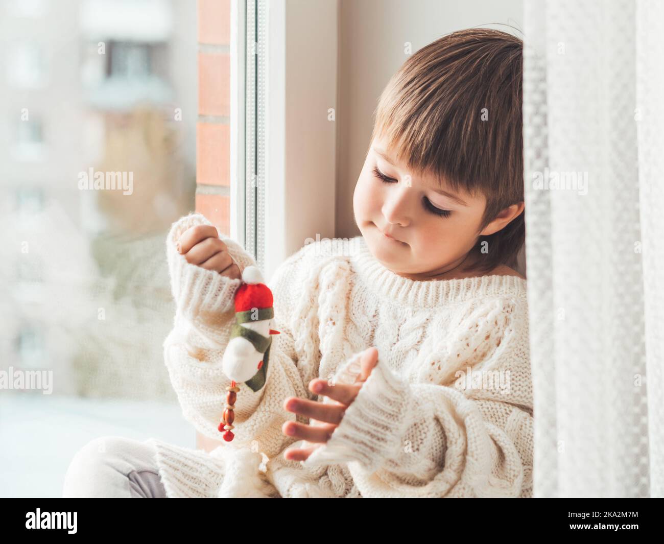 Kid with felt decorative snowman for Christmas tree. Boy in cable-knit oversized sweater. Cozy outfit for snuggle weather. Character with Santa hat. Stock Photo