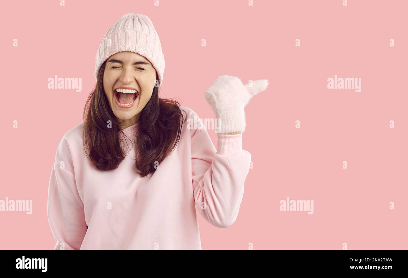 Funny woman in winter clothes laughing while pointing to copy space side on pink background Stock Photo