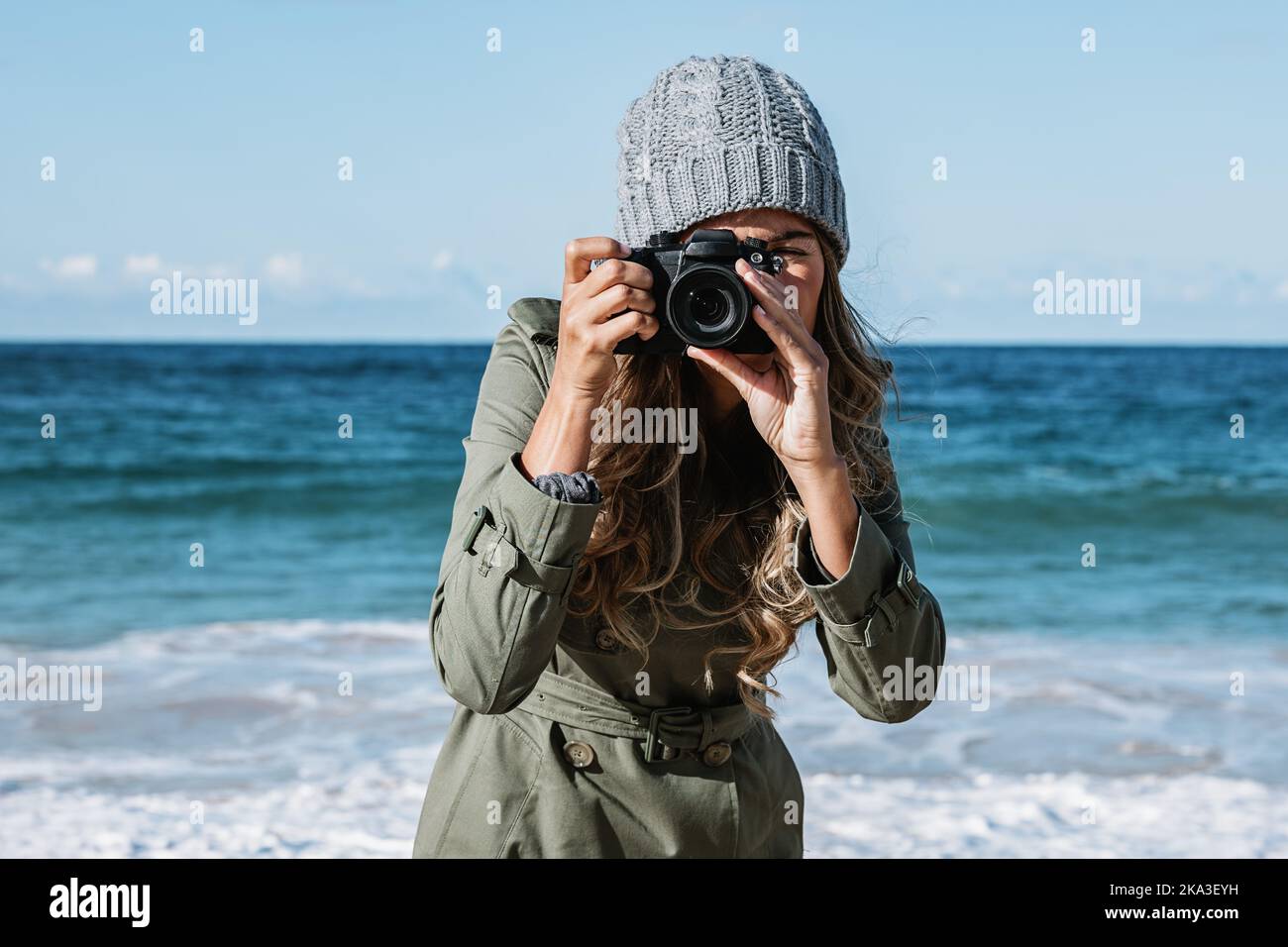 Female photographer in knitted hat and trench coat shooting at camera